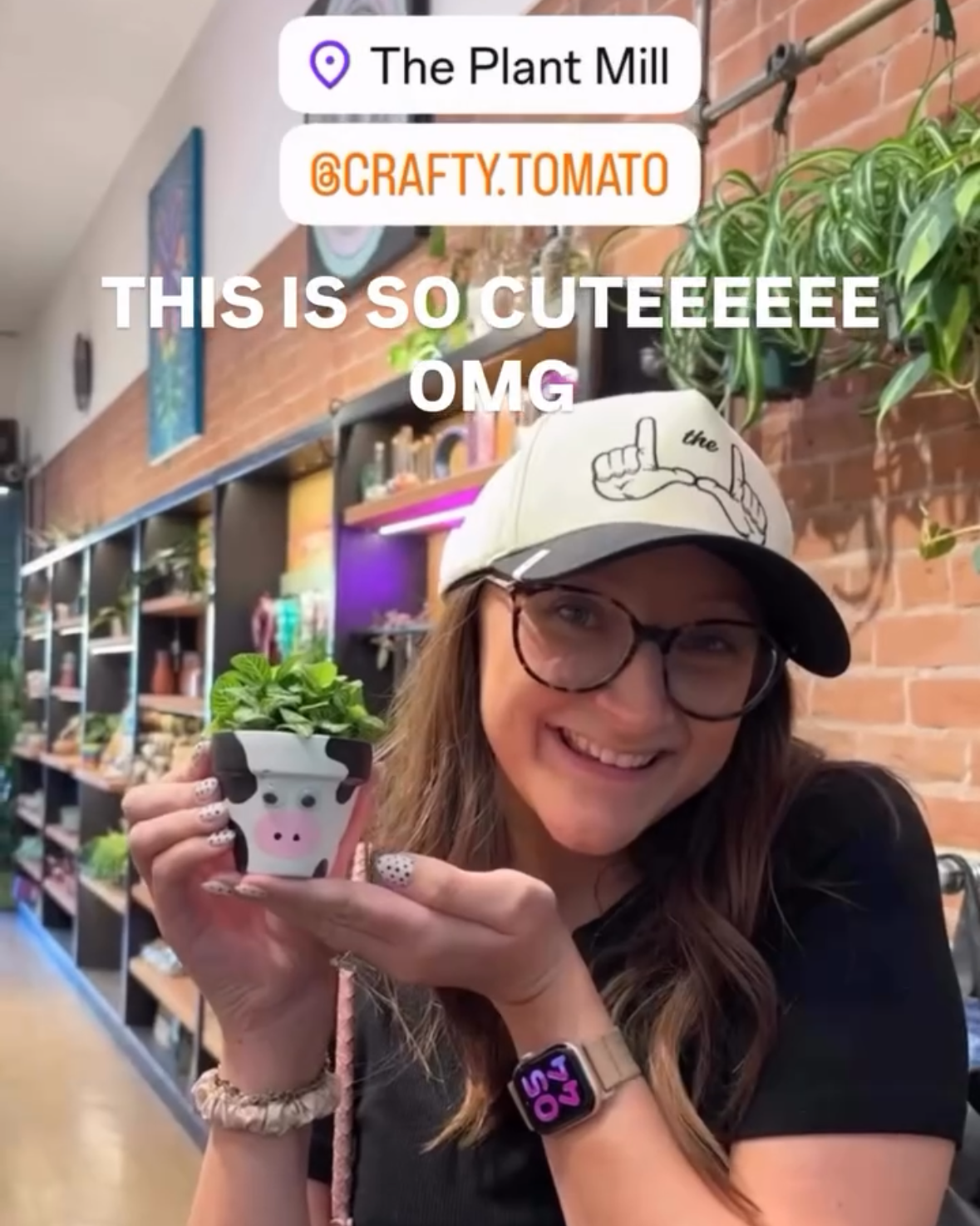 A woman wearing a white baseball cap and glasses holds up a small potted cow plant in a shop setting. The image has a phone screen overlay showing location and social media text.