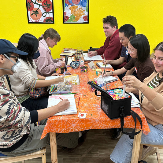 A group of people sitting around a table in a brightly lit room with a yellow wall, working on art or writing activities. The table has an orange spiderweb-patterned tablecloth, and several colorful art supplies and books are visible on it.