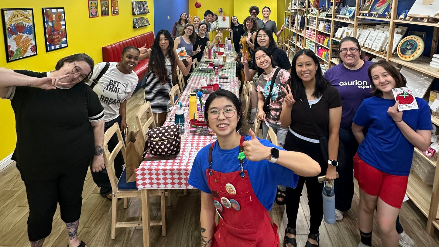 A group of people posing together in a colorful indoor shop or classroom setting with yellow walls, wooden floors, and shelves filled with items. The central person is wearing a bright red apron over a blue shirt and giving a thumbs-up.