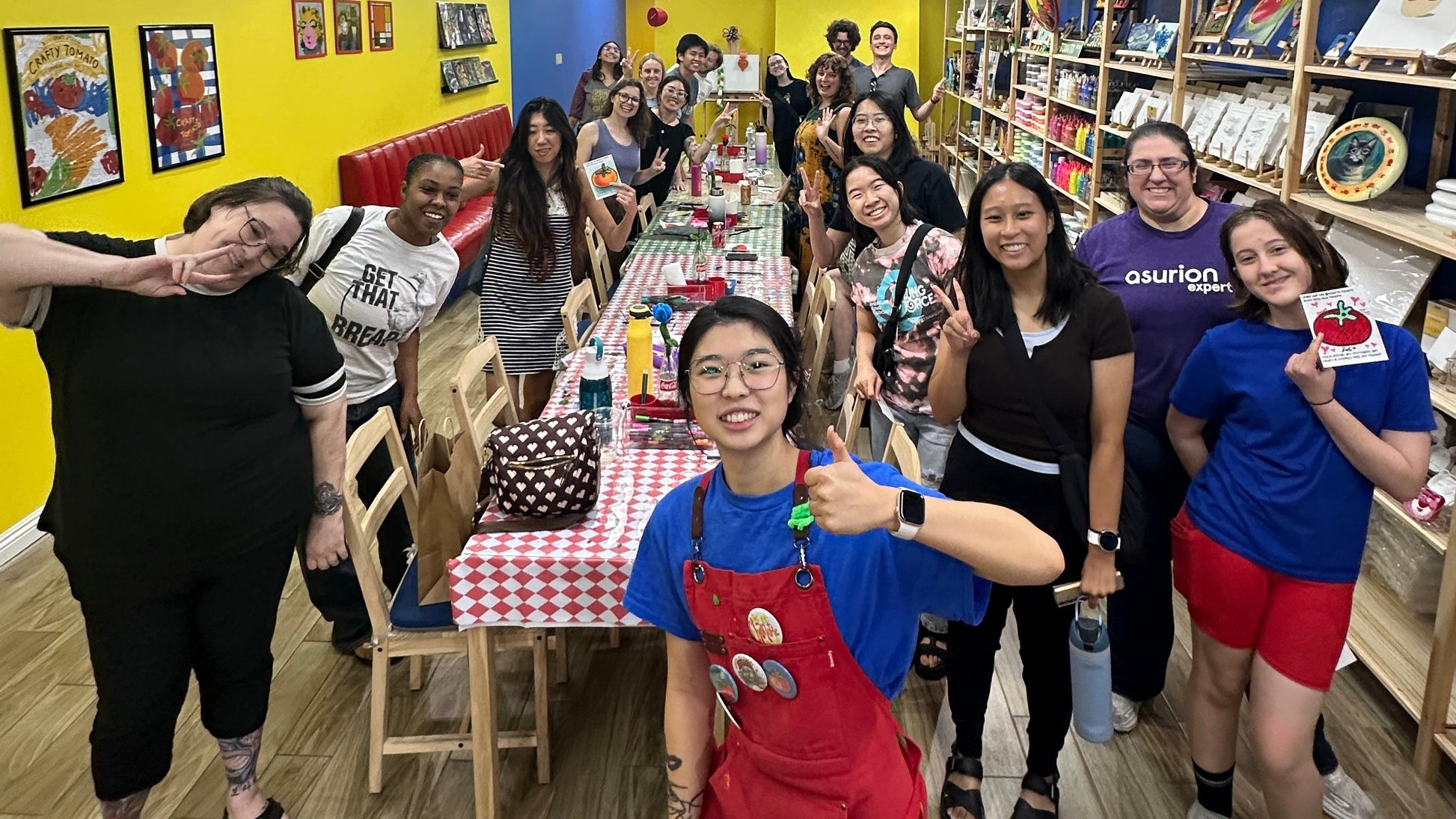 A group of people posing together in a colorful indoor shop or classroom setting with yellow walls, wooden floors, and shelves filled with items. The central person is wearing a bright red apron over a blue shirt and giving a thumbs-up.