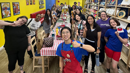 A group of people posing together in a colorful indoor shop or classroom setting with yellow walls, wooden floors, and shelves filled with items. The central person is wearing a bright red apron over a blue shirt and giving a thumbs-up.