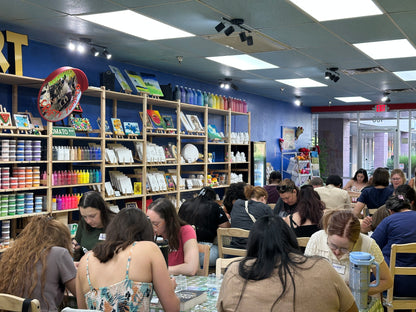 A busy art studio or craft shop interior with multiple people seated at tables, shelves filled with colorful art supplies, and blue walls in the background.