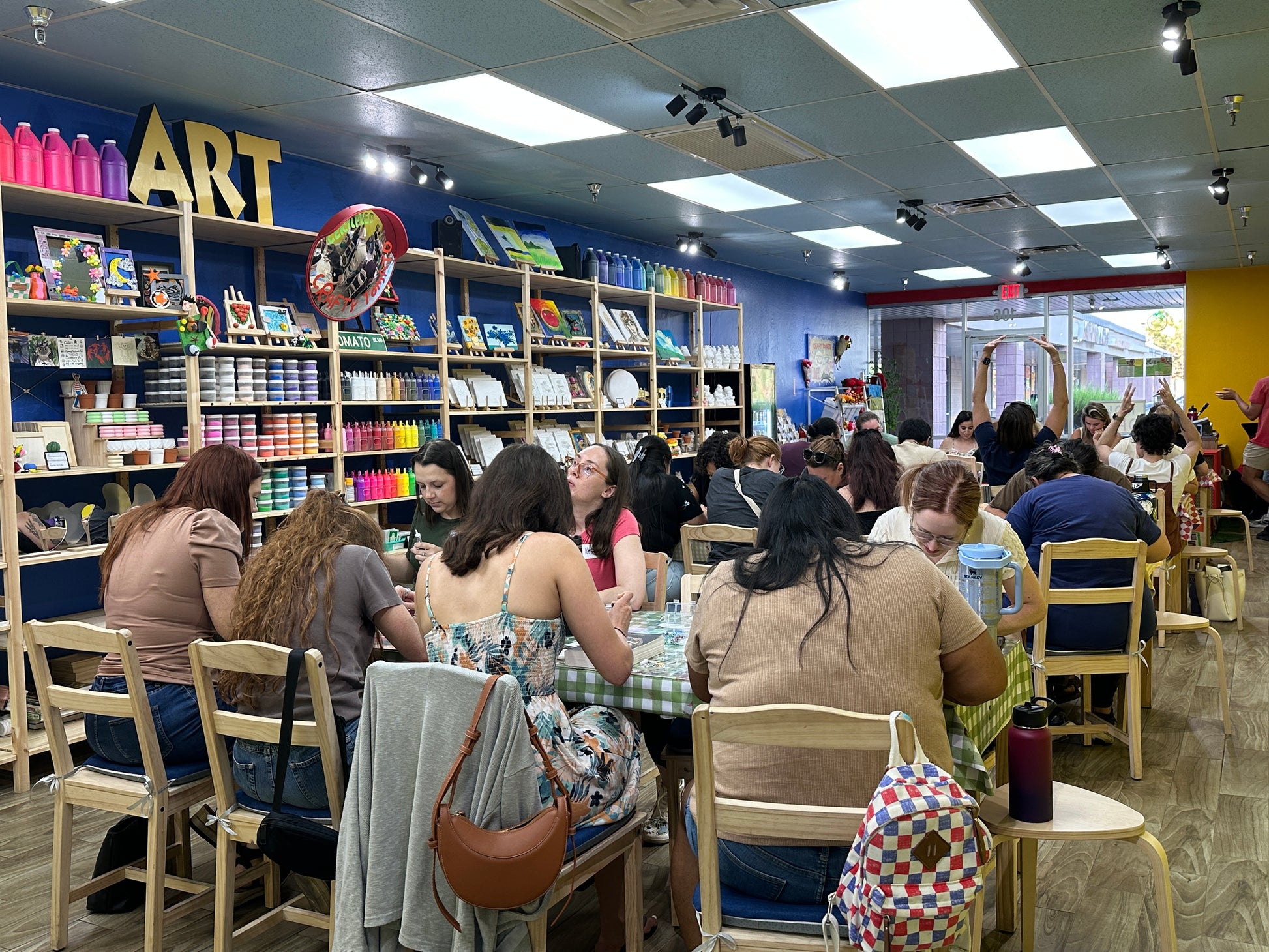 A busy art studio or classroom filled with people seated at tables, surrounded by shelves of colorful art supplies and paint bottles against a blue wall with a large yellow 'ART' sign.