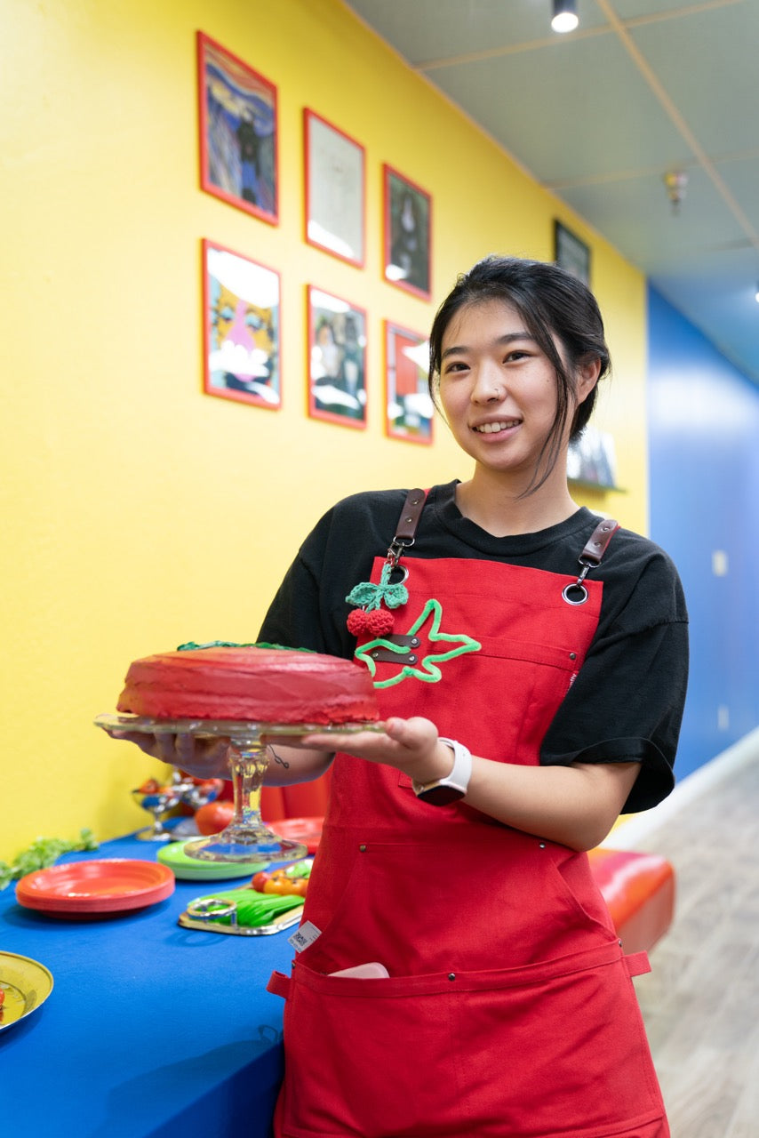 Person in a red apron holding a cake in a colorful room with a yellow wall and blue table.