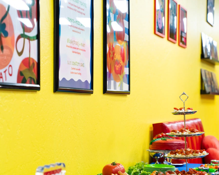 Buffet setup with drinks, vegetables, and a colorful wall with framed pictures.