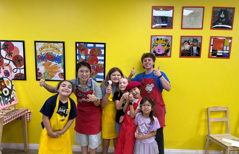 A group of art teachers and children wearing colorful aprons and chef hats pose together in a bright yellow room with framed artwork on the wall.
