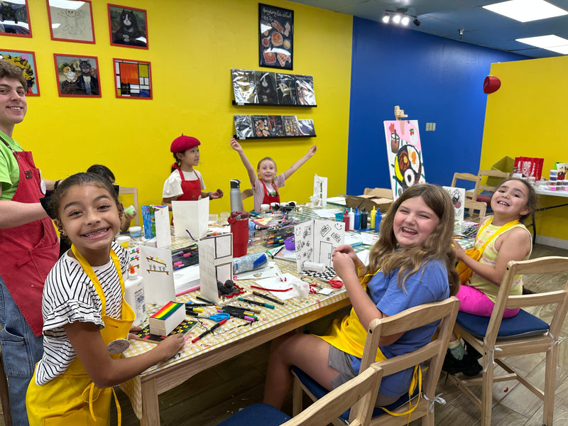 A group of children smiling and painting at a table in a colorful craft studio with yellow and blue walls.