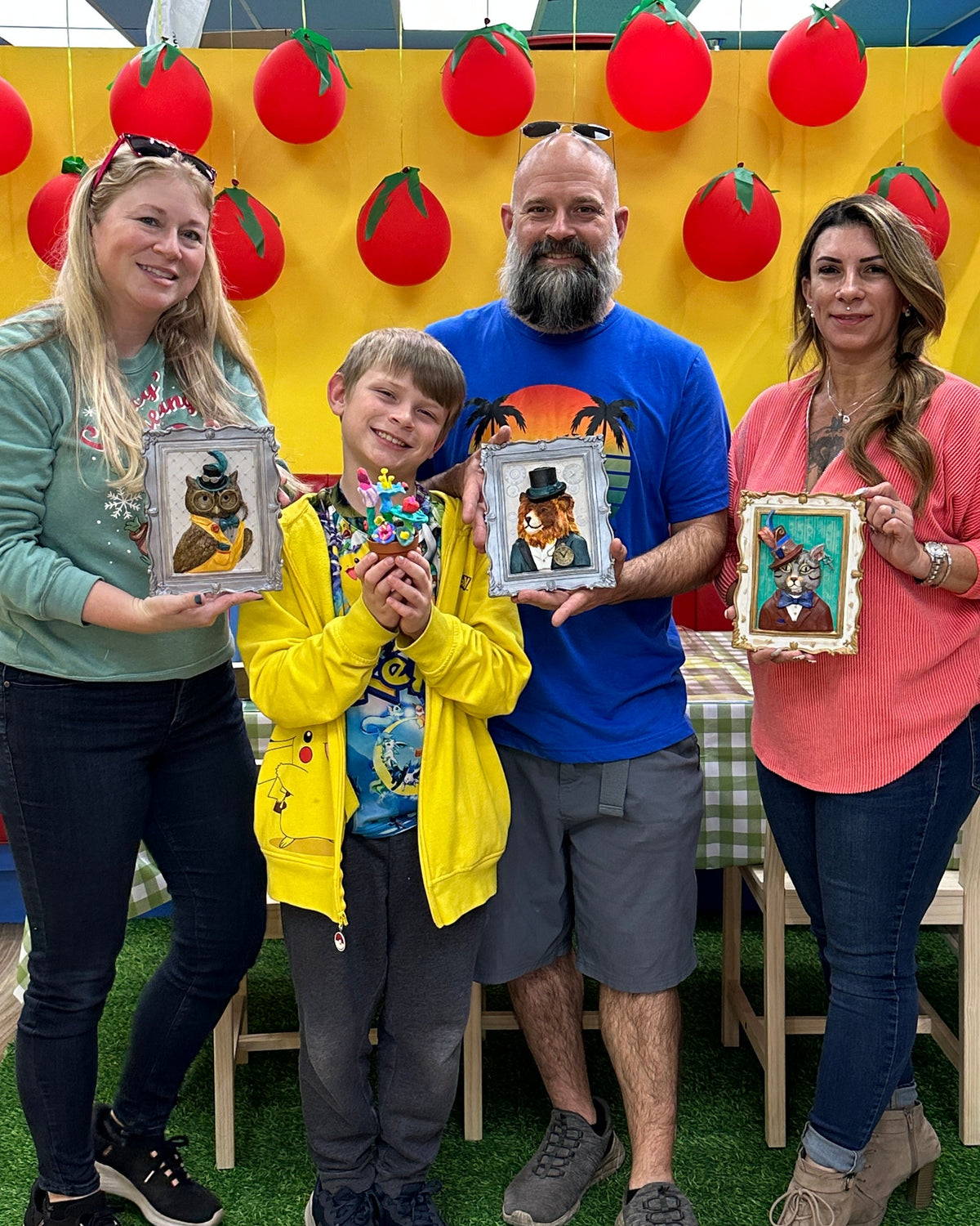 A family of four poses together in a colorful indoor playroom or daycare setting, each holding a small framed picture. The background features a bright yellow wall decorated with red tomato shapes, blue side walls, and a green artificial turf floor.