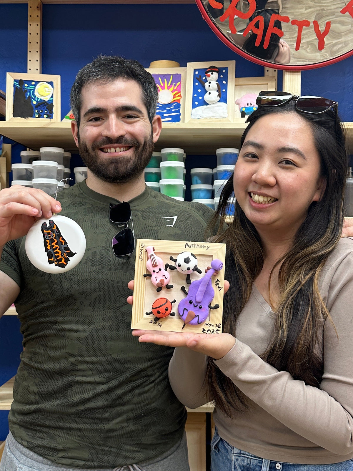 A man and a woman are standing in a colorful craft or art studio, smiling and holding a small illustrated card or print. The background shows shelves filled with colorful supplies, bottles, and framed items against a blue wall.
