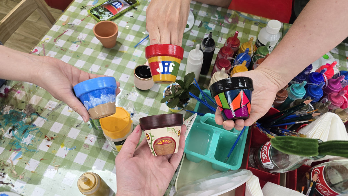 A group of people holding small, colorful painted flower pots over a paint-splattered green and white checkered table covered with paint bottles and brushes.