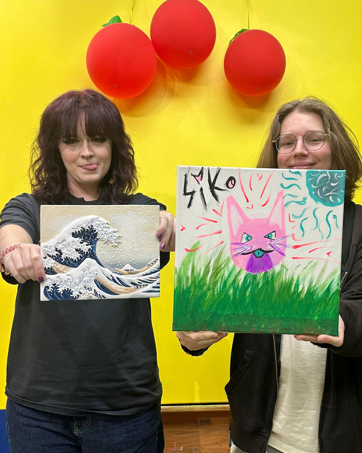 Two women standing indoors in front of a large yellow backdrop with red balloons, each holding up a painted canvas. The woman on the left holds a blue-gray painting of a wave scene, and the woman on the right holds a colorful painting with a pink bunny, green grass, and the word 'Sko'.