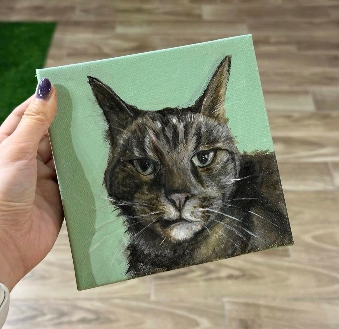 Hand holding a small square canvas with a cat painting on a wooden floor.