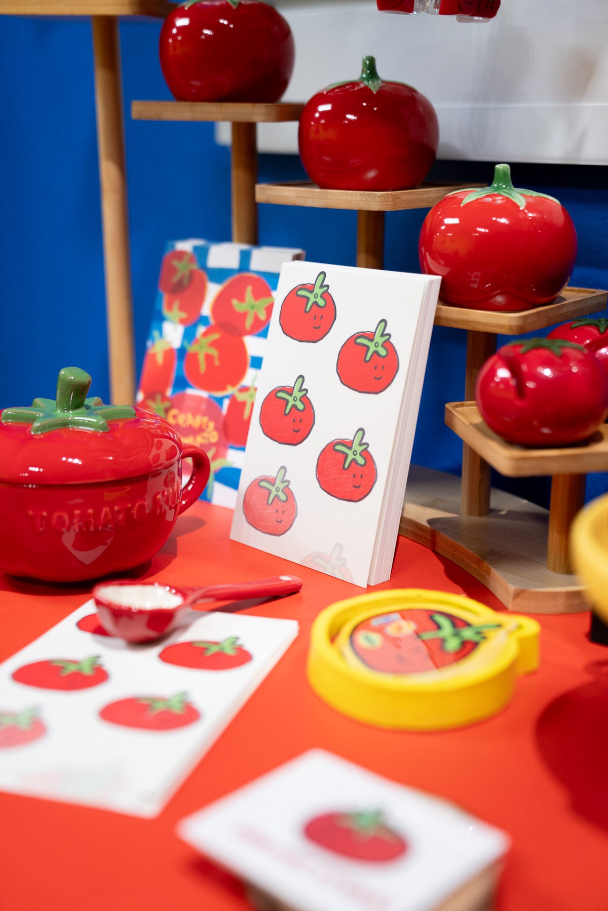 A children's toy play kitchen set featuring red ceramic tomato-shaped pots, a yellow lid, and printed cards with tomato illustrations, arranged on a red table with wooden display shelves in the background.
